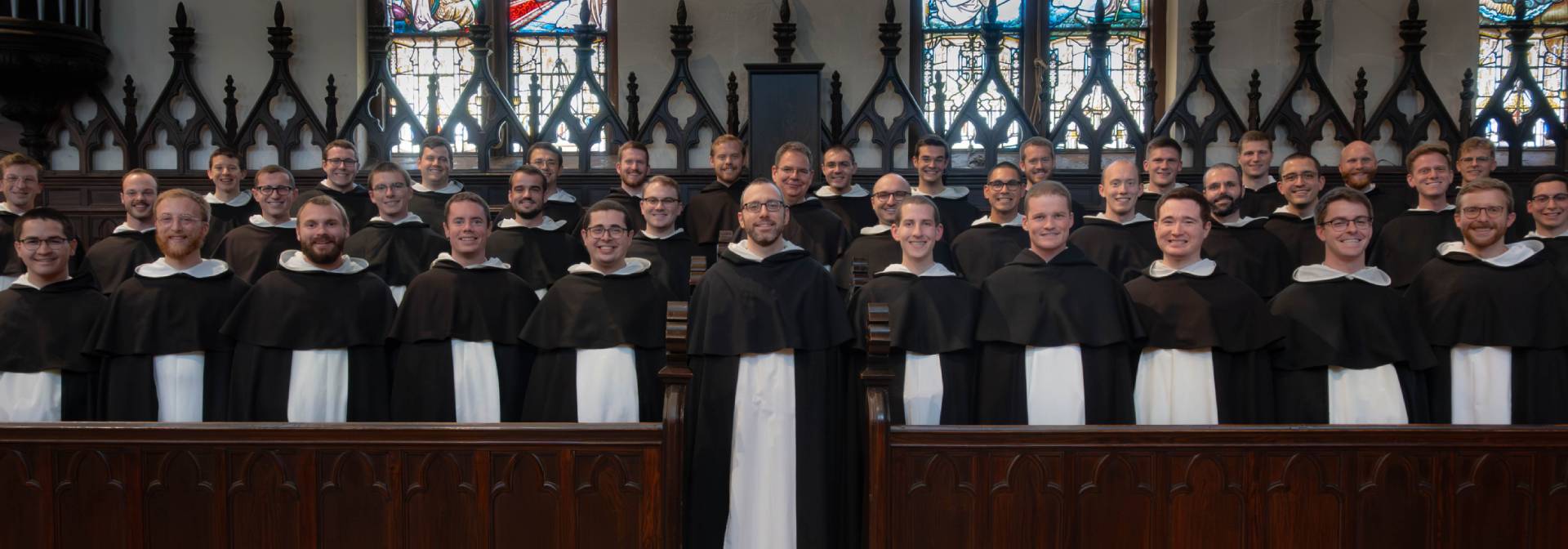 A group of men in dark robes and white collars standing in a church setting with stained glass windows in the background.